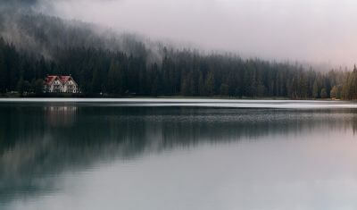 A beautiful cottage sits on the edge of a wooded hillside, seen from across a foggy lake on a misty day.