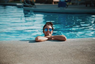 Smiling girl wearing sunglasses is inside a swimming pool leaning against the side.